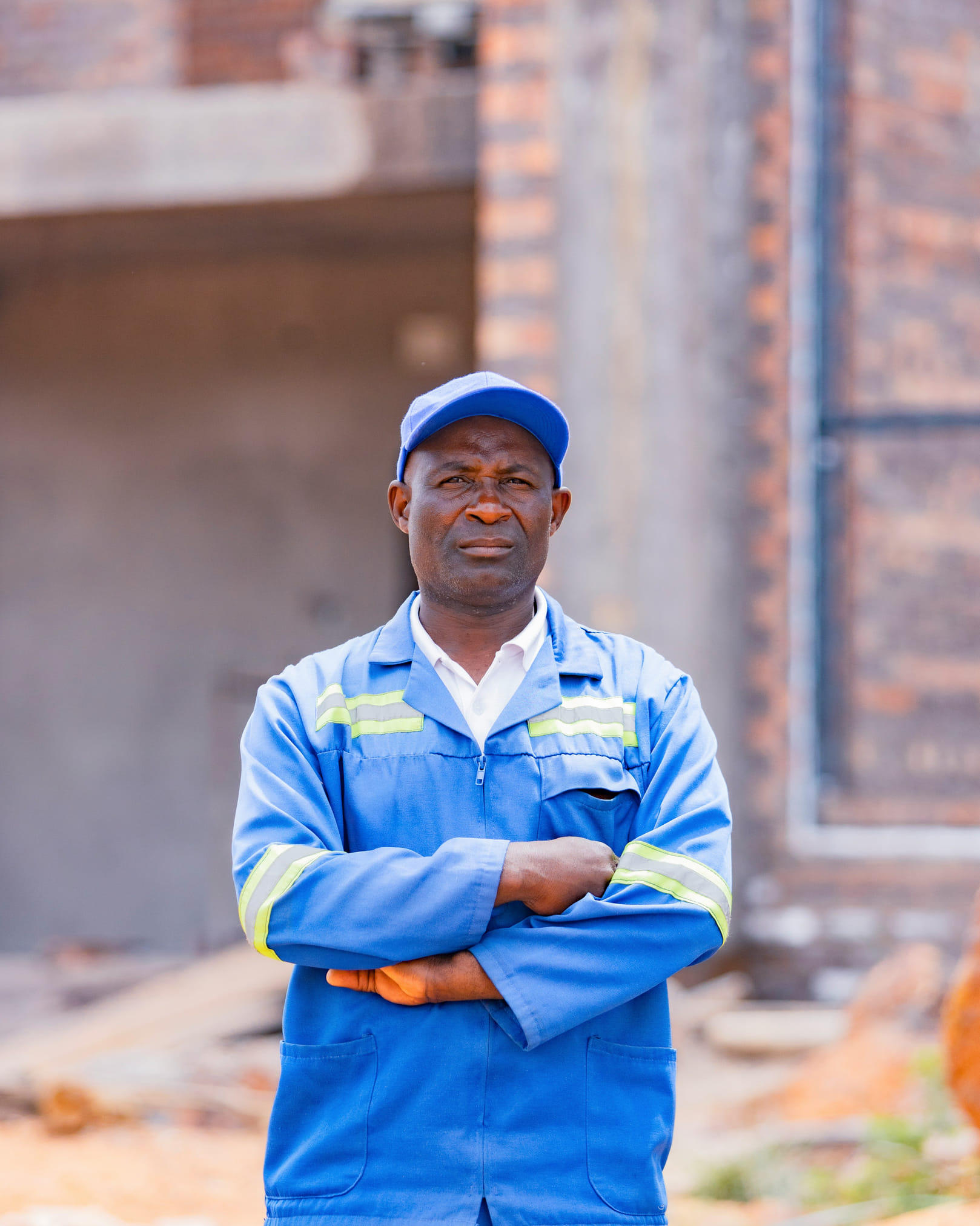 A tradesperson in blue coveralls with reflective stripes and a blue cap, arms folded, standing in front of an unfinished brick building.