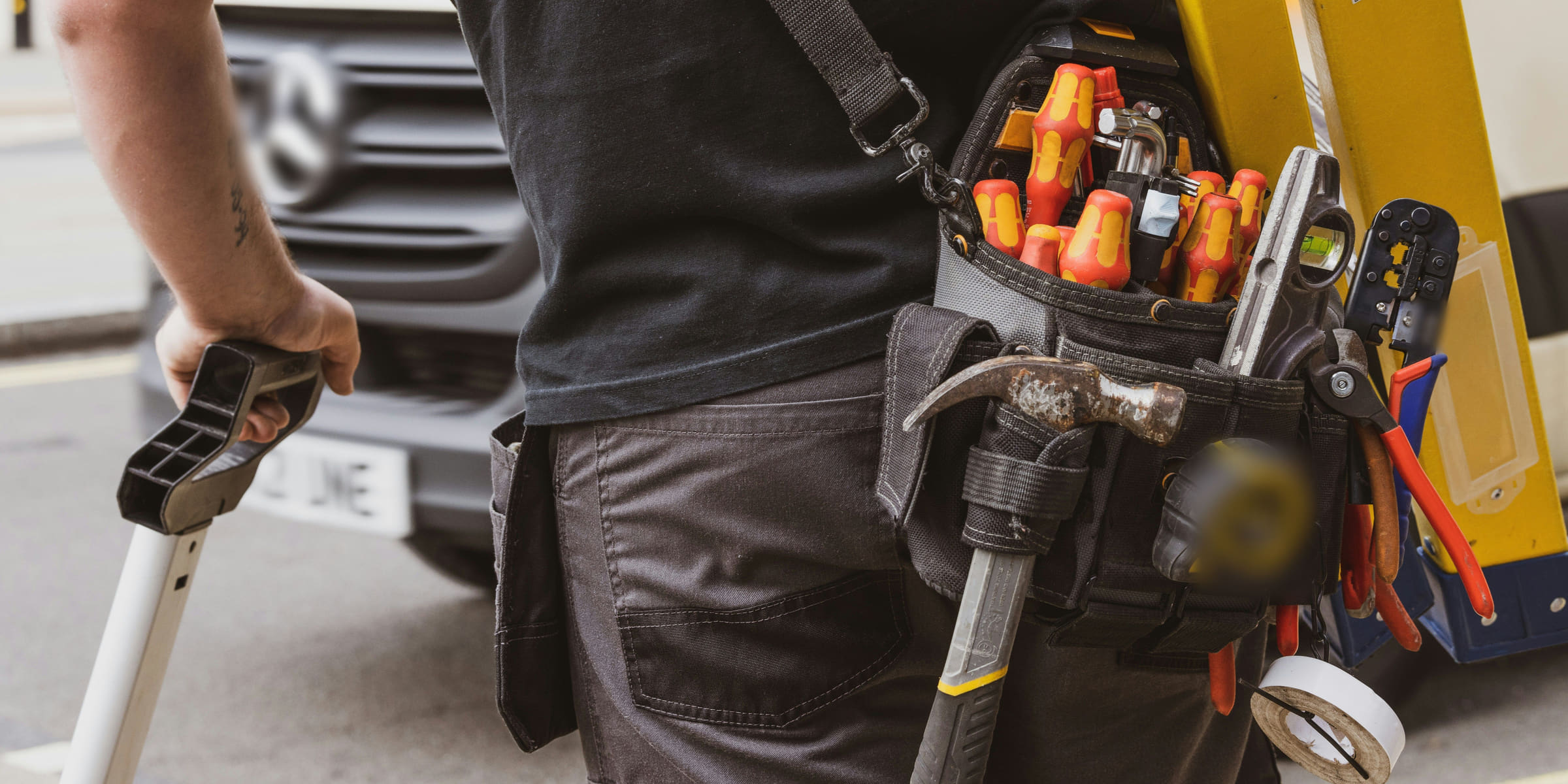 A technician seen from behind carrying a fibreglass step ladder, wearing a fully loaded tool belt of screwdrivers, pliers and a hammer, beside a parked work van.