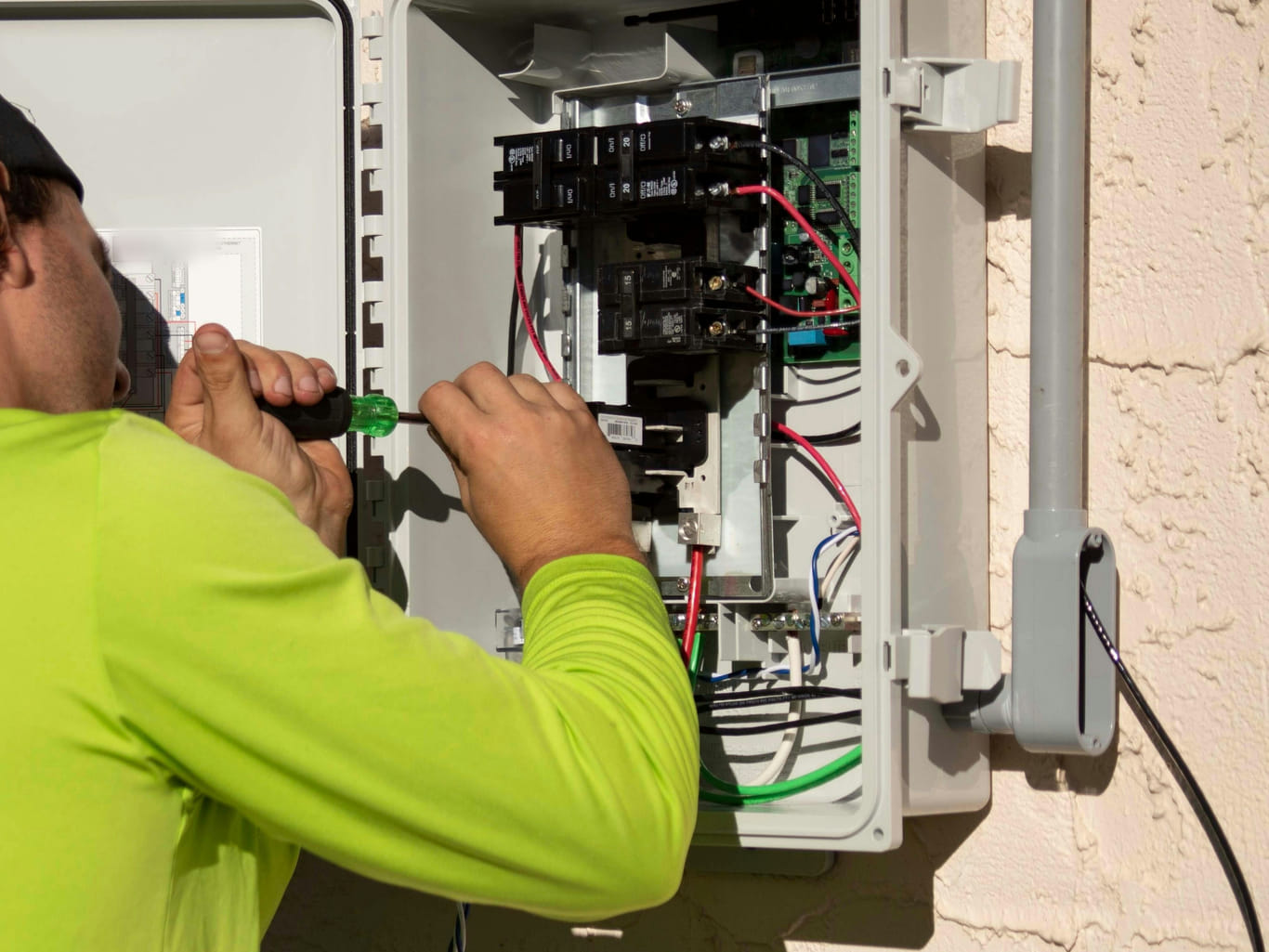 A technician's hands working with a screwdriver inside an open outdoor electrical enclosure, with breakers and coloured wiring visible.