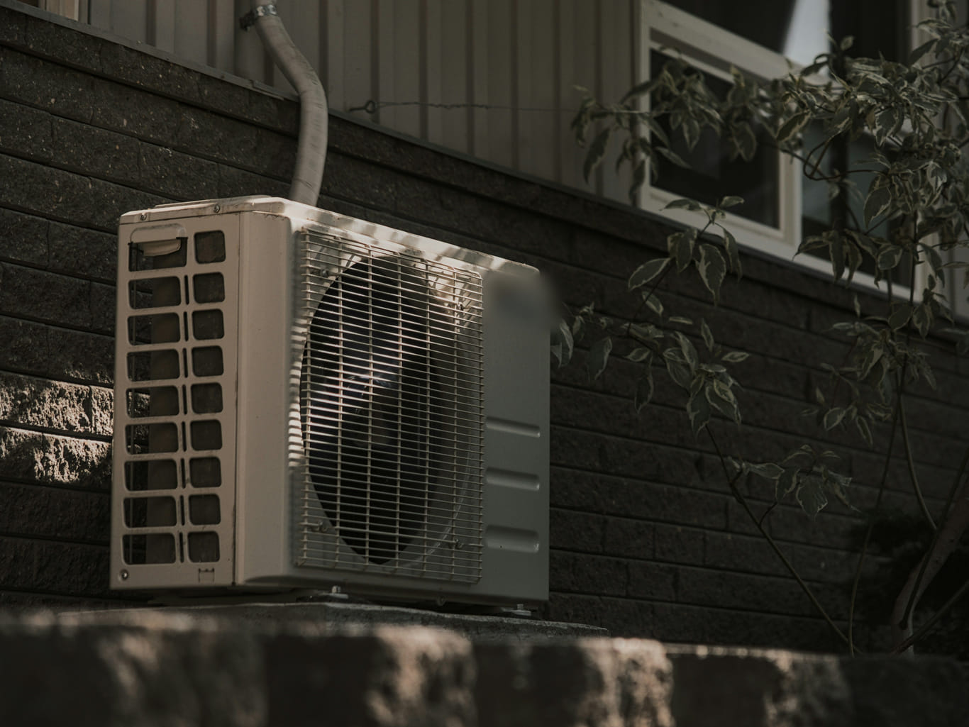 An outdoor air conditioning condenser unit mounted against a dark brick wall in low afternoon light.