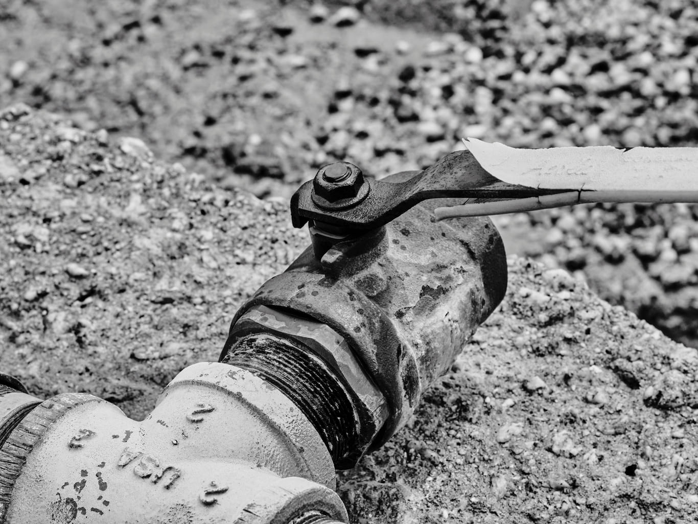 Black and white close-up of a cast iron ball valve and threaded pipe fitting on an exposed water line running through gravel.
