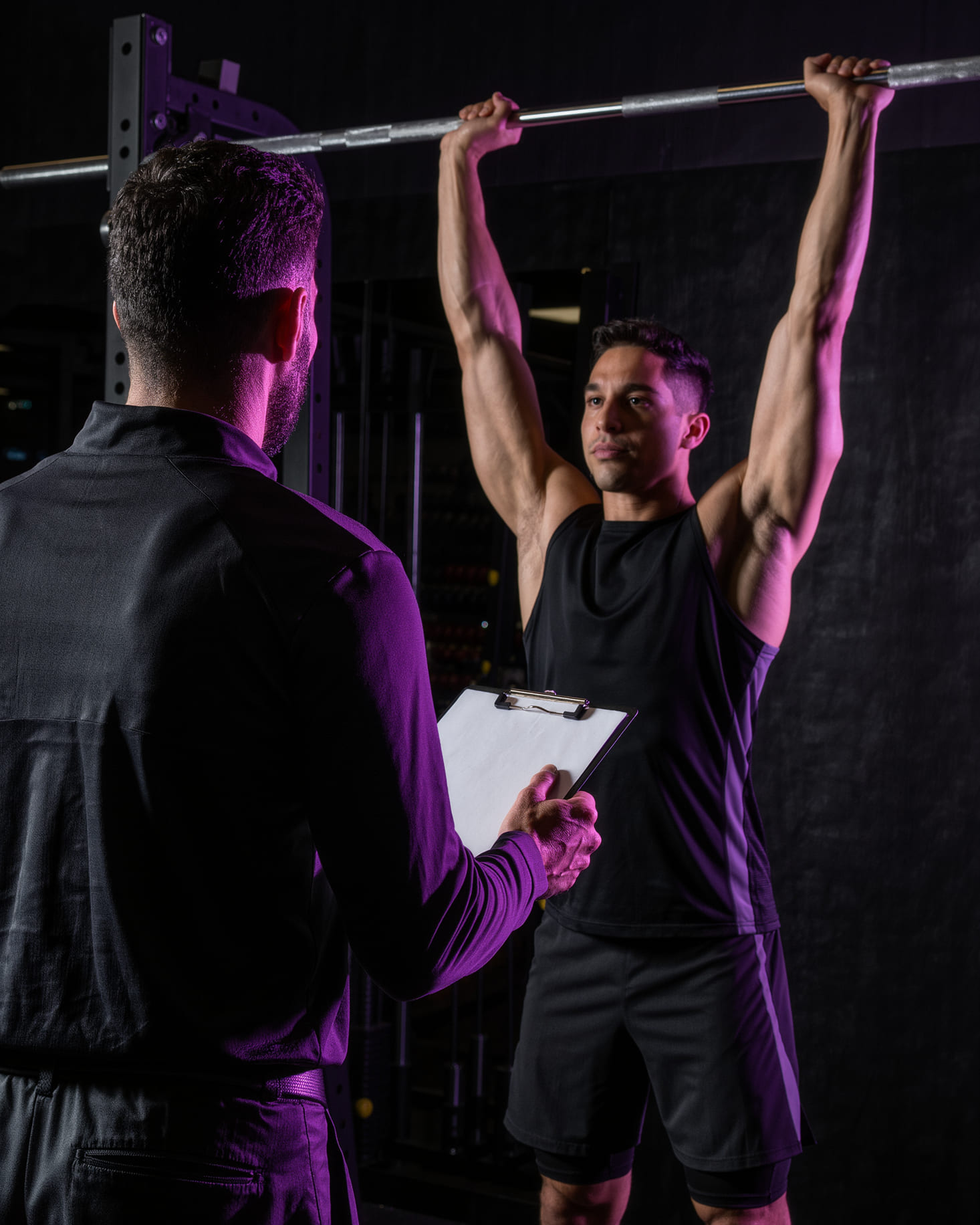 A coach stands with his back to the camera holding a clipboard, watching a client hold a barbell overhead at lockout under violet gym lighting.