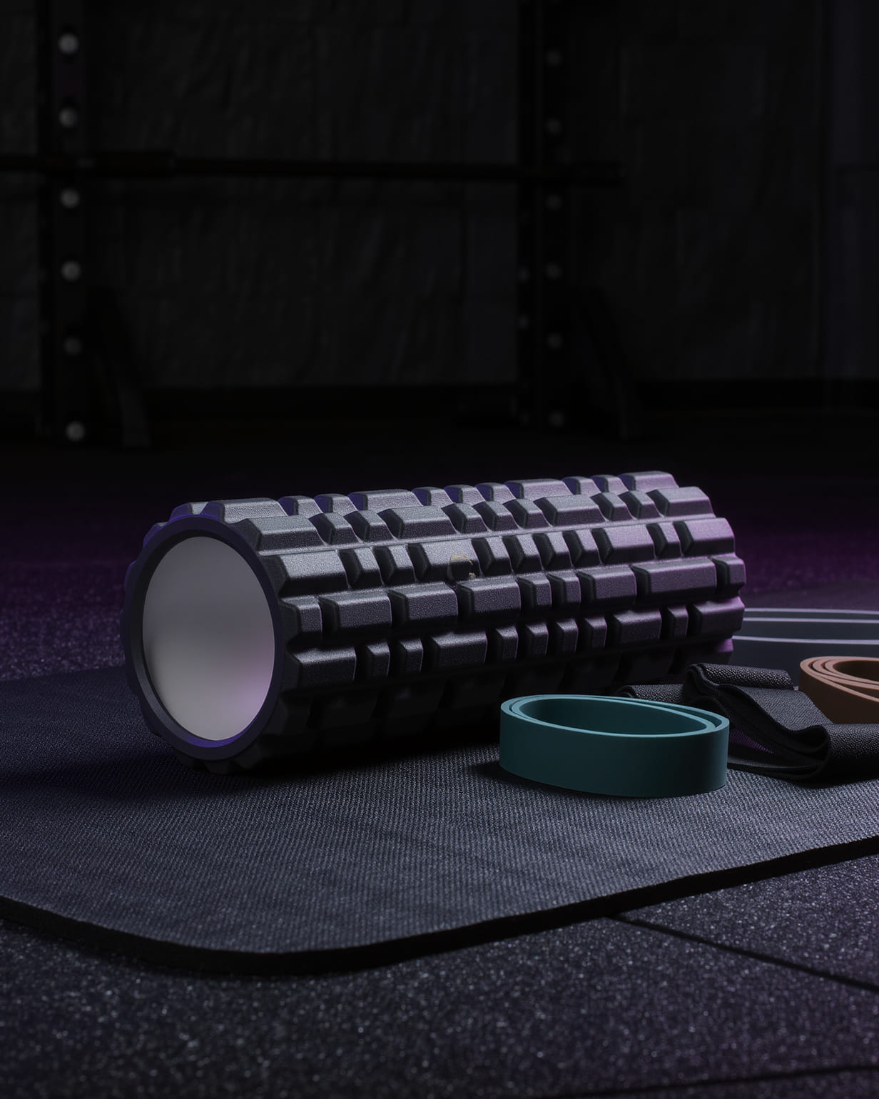 A textured foam roller lying on a mat beside looped resistance bands, a squat rack out of focus behind them.