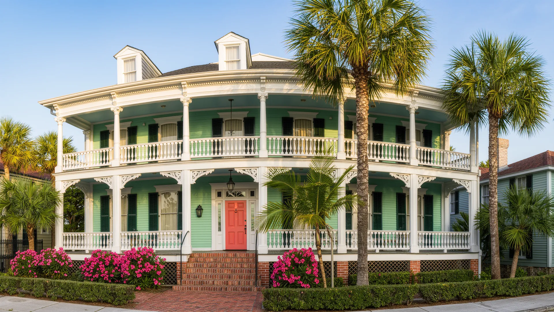 A two story Victorian house painted pale sea green with white double gallery porches, a coral front door, palm trees and pink flowers in the garden