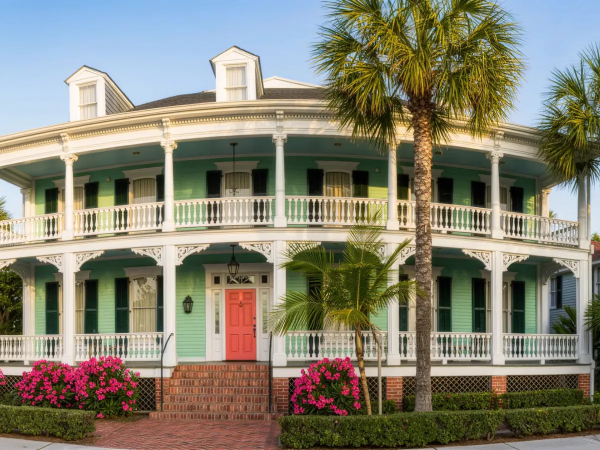A two story Victorian house painted pale sea green with white double gallery porches, a coral front door, palm trees and pink flowers in the garden