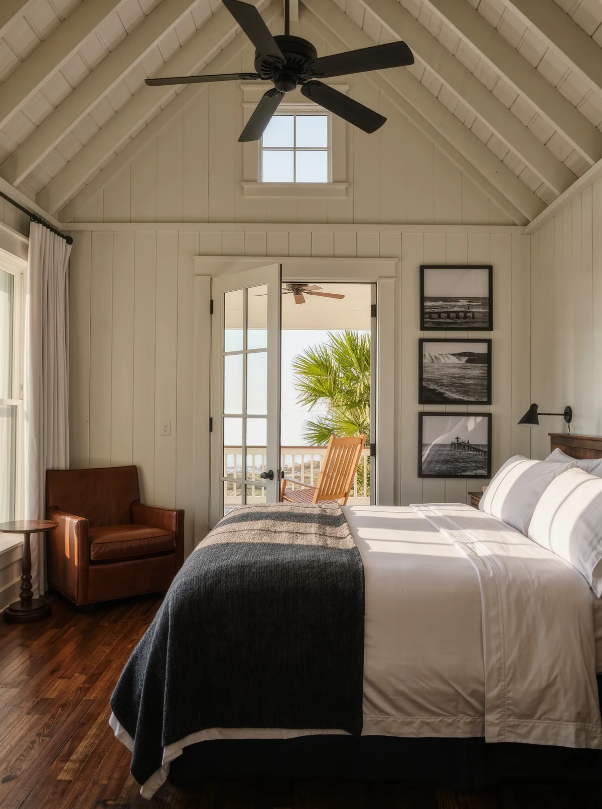 A carriage house suite with a vaulted board ceiling, a king bed with a charcoal blanket, black and white photographs on the wall, a leather armchair and French doors open to a porch