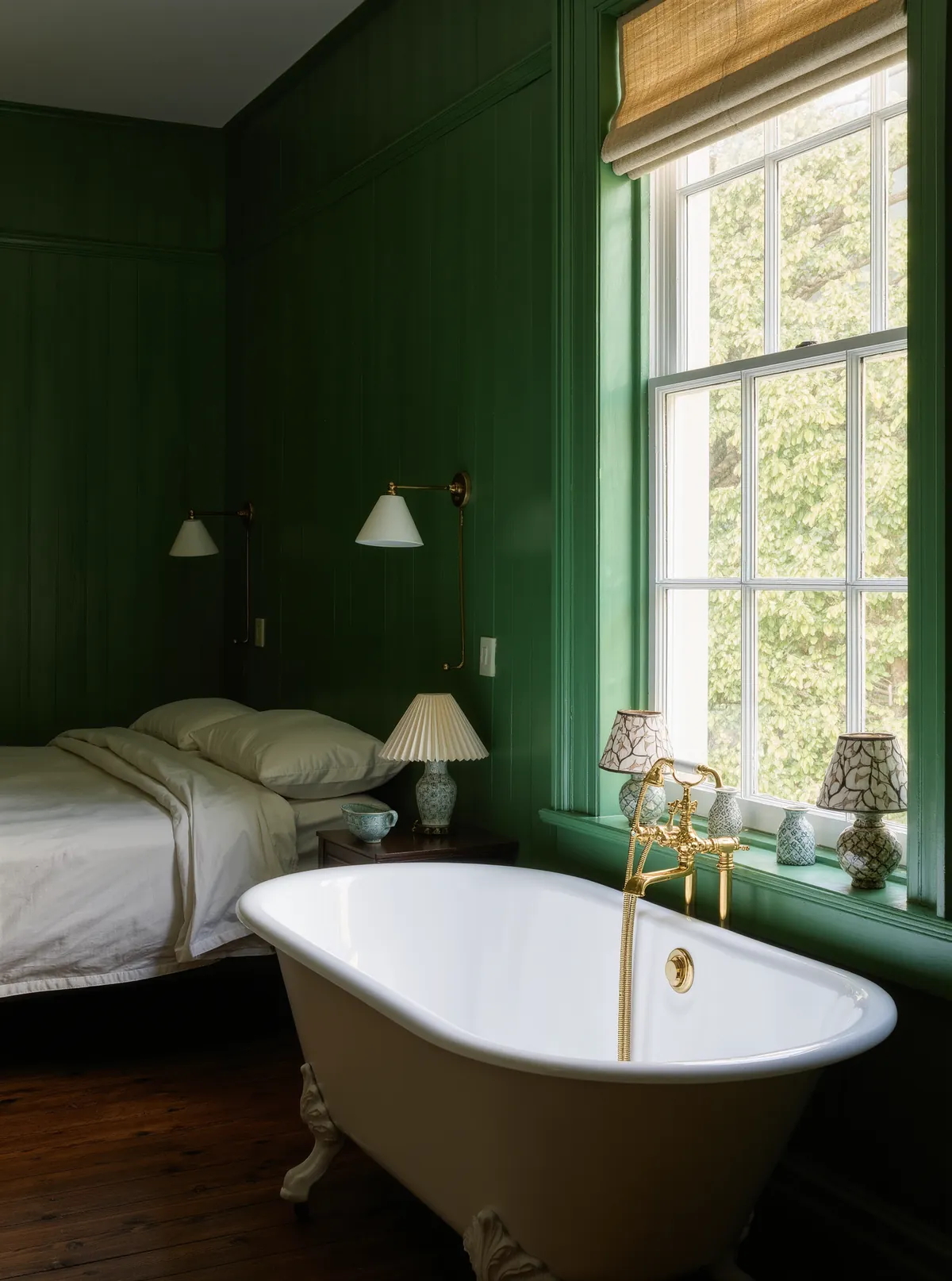 A guest room with deep green walls, a white clawfoot tub with brass taps under a tall window and a bed in the background