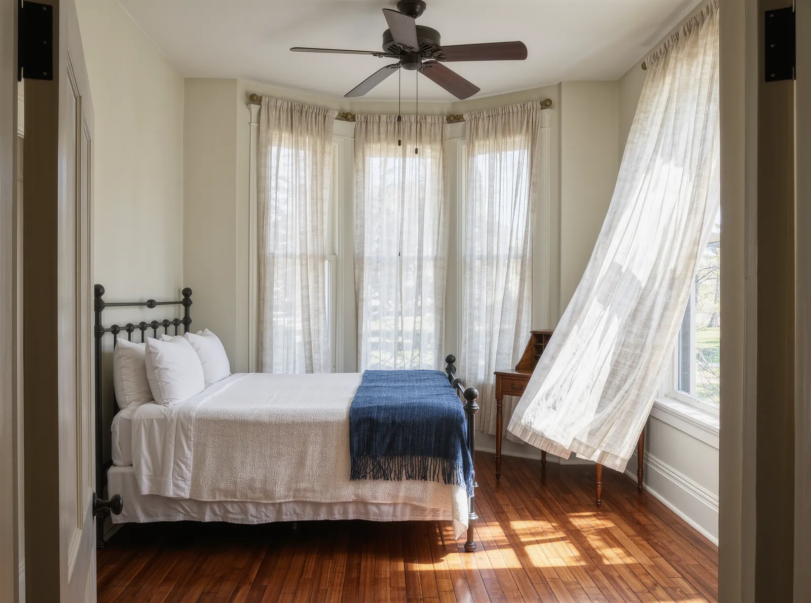 A bright corner bedroom with a black iron bed, white linen, an indigo throw and sheer curtains lifting at tall windows