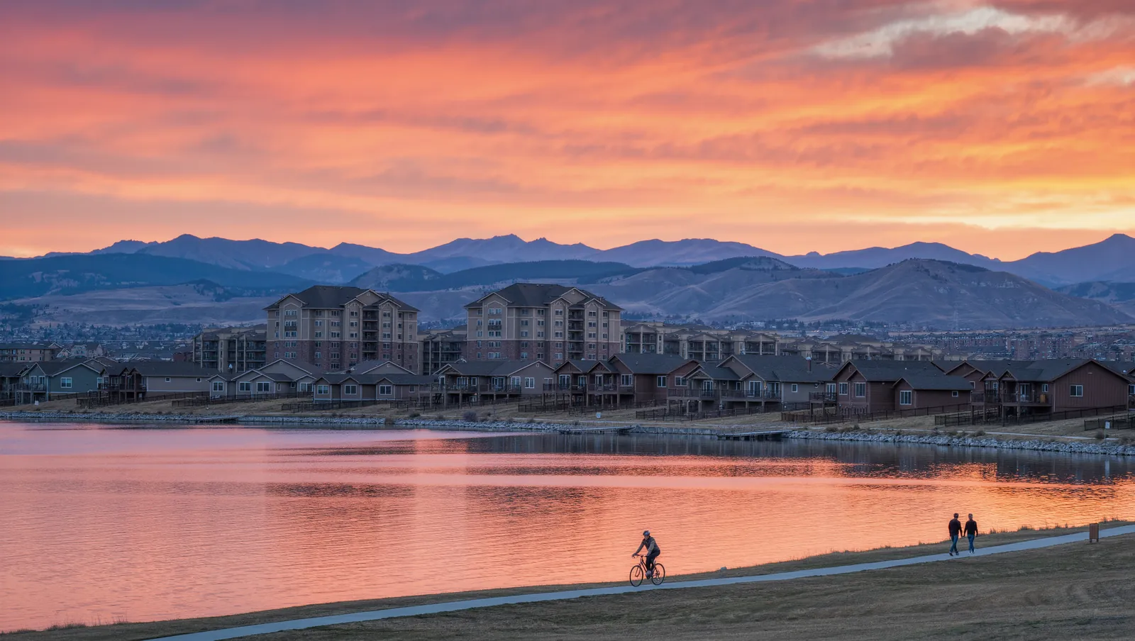 A wide city lake at sunset with a cyclist on the shore path and the Front Range mountains behind