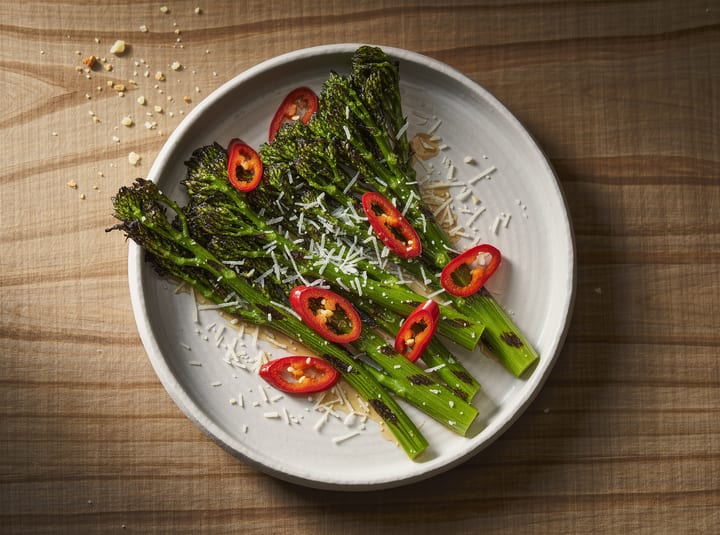 Charred broccolini spears on a pale plate, scattered with sliced red Calabrian chili and shaved parmesan.