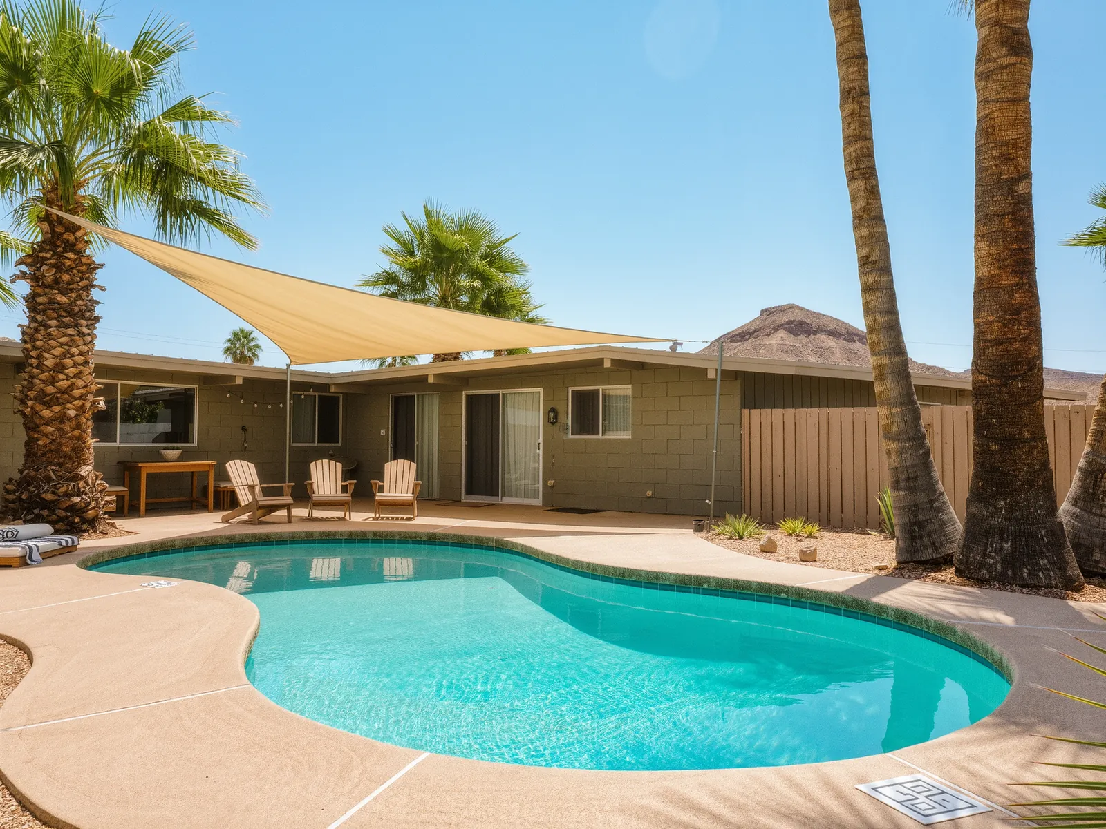 A small kidney shaped pool behind a mid century block home with tall palm trees and a shade sail